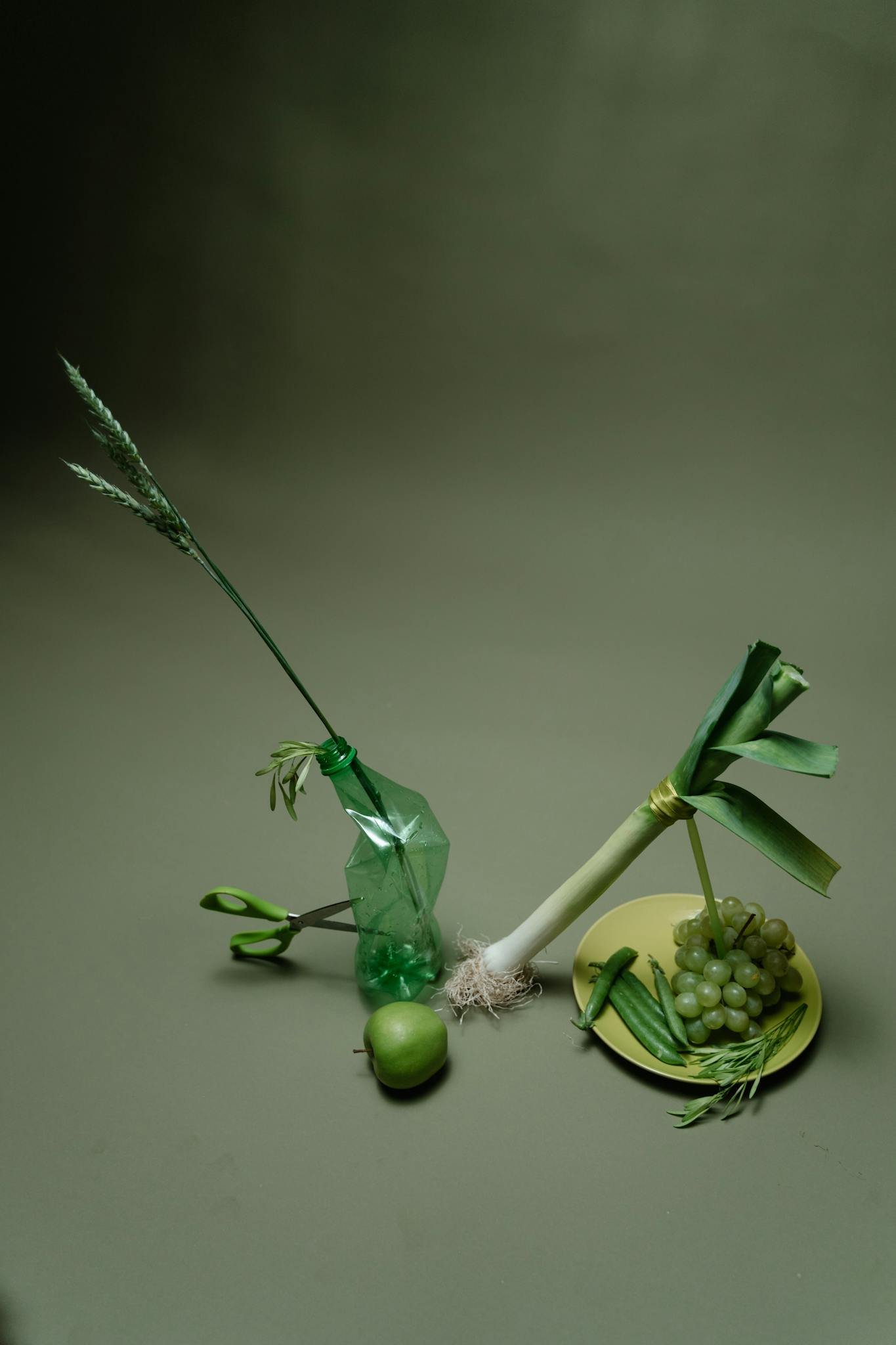 Artistic arrangement of green vegetables, fruits, and a bottle in a still life photo.